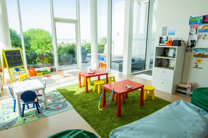 A bright children's playroom featuring colorful tables and chairs, a carpet, toys, and large windows allowing natural light to fill the space.