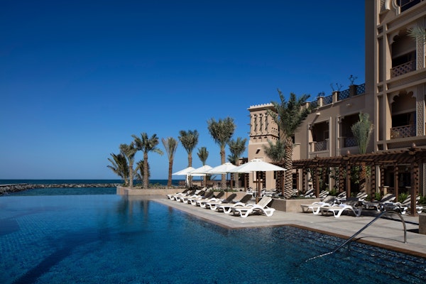 A luxurious poolside view at a resort with white lounge chairs, palm trees, and a clear blue sky overlooking the ocean.