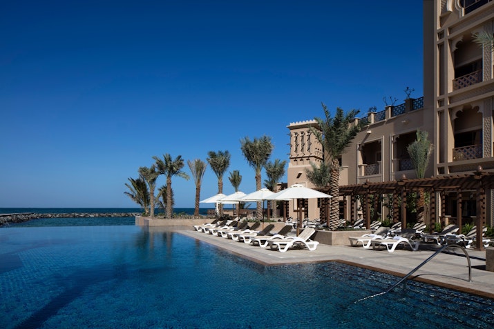 A luxurious poolside view at a resort with white lounge chairs, palm trees, and a clear blue sky overlooking the ocean.