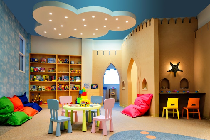A colorful children's playroom featuring bean bags, a play table with chairs, and shelves filled with toys under a whimsical cloud-shaped ceiling.