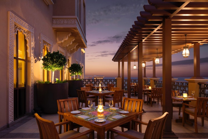A cozy outdoor restaurant setting illuminated by warm lights at dusk, featuring wooden tables and lush greenery with an ocean view in the background.