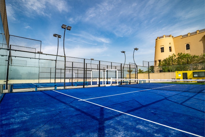 A blue paddle tennis court with glass walls and lighting fixtures, set against a clear sky and a historic building in the background.