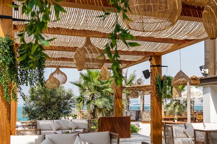 A beachside seating area under a woven canopy, with hanging lights and tropical plants surrounding the space.