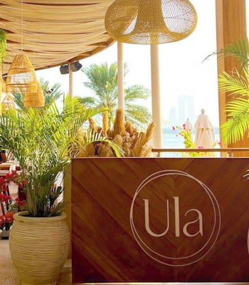 Ula Dubai Restaurant featuring wooden decor, lush indoor plants, and large windows showcasing a view of the outdoors.