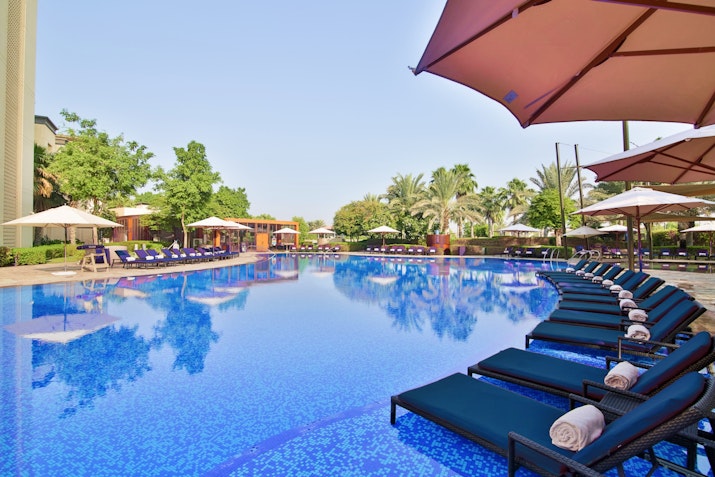 Large pool with blue loungers, umbrellas, and surrounding greenery.