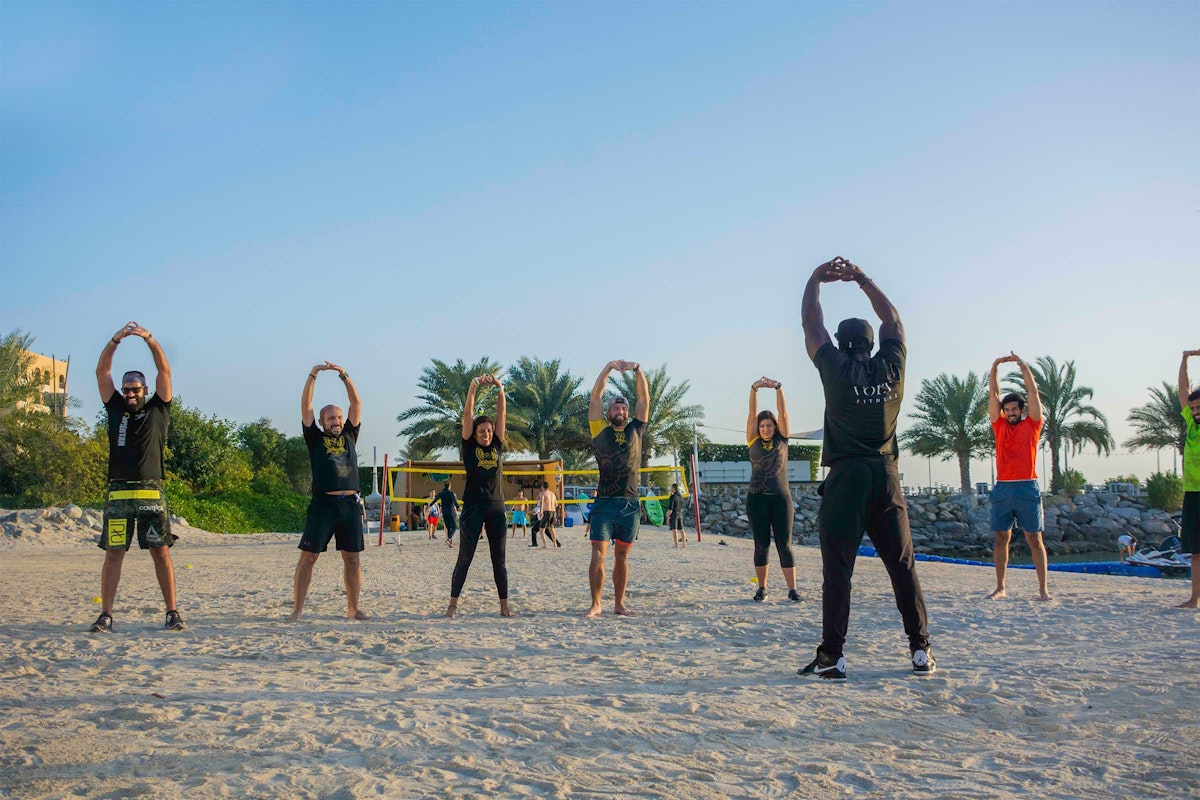 A group of people exercising on the beach, stretching their arms in the air, with palm trees in the background.