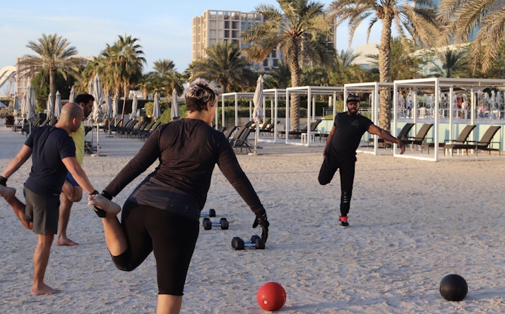 A group of people performing a warm-up exercise on the beach, with weights nearby and palm trees in the background.