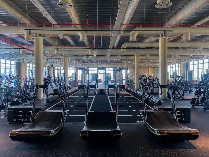 Wide gym space filled with advanced treadmills and fitness machines, featuring high ceilings and industrial-style lighting.