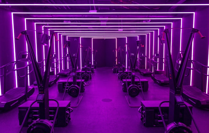 Modern gym cardio zone with neon purple lighting outlining the walls and ceiling, featuring treadmills and rowers in a striking, futuristic setting.