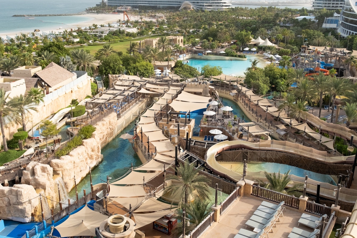 Aerial view of Wild Wadi Waterpark featuring winding water slides, pools, shaded lounging areas, and lush palm trees, with the Arabian Gulf and beachfront hotels in the background
