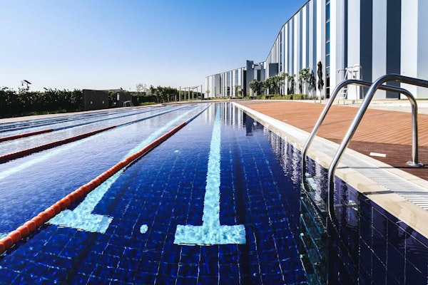 Outdoor Olympic-style swimming pool with lane dividers, reflecting the bright blue sky at WellFit Meydan in Dubai, flanked by modern architecture and a pool deck.