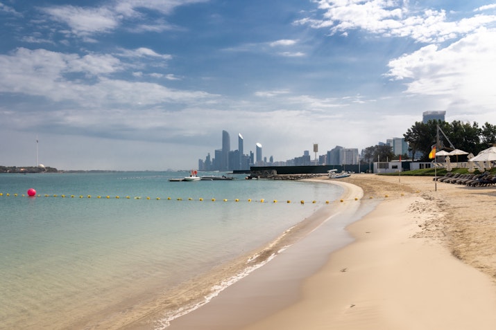 Sandy beach with calm water, city skyline, and cloudy sky in the background.
