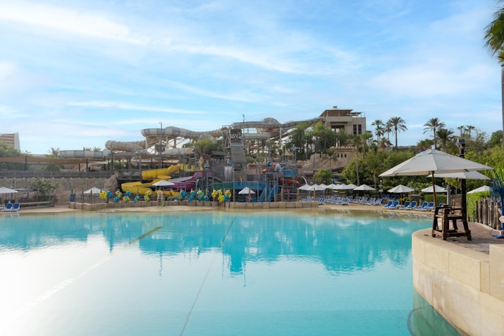 A large, calm pool with clear blue water, surrounded by sun loungers and umbrellas, with various water slides and tropical greenery visible in the background under a bright sky.