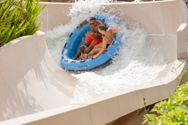 A man and woman enjoying a thrilling ride on a blue inflatable raft down a water slide, surrounded by rushing water and lush green plants