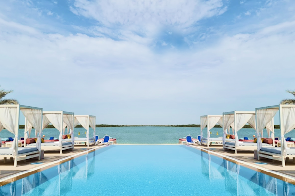 Yas Beach, Abu Dhabi A serene infinity pool with lounge beds and canopies overlooking calm waters and a blue sky with wispy clouds.
