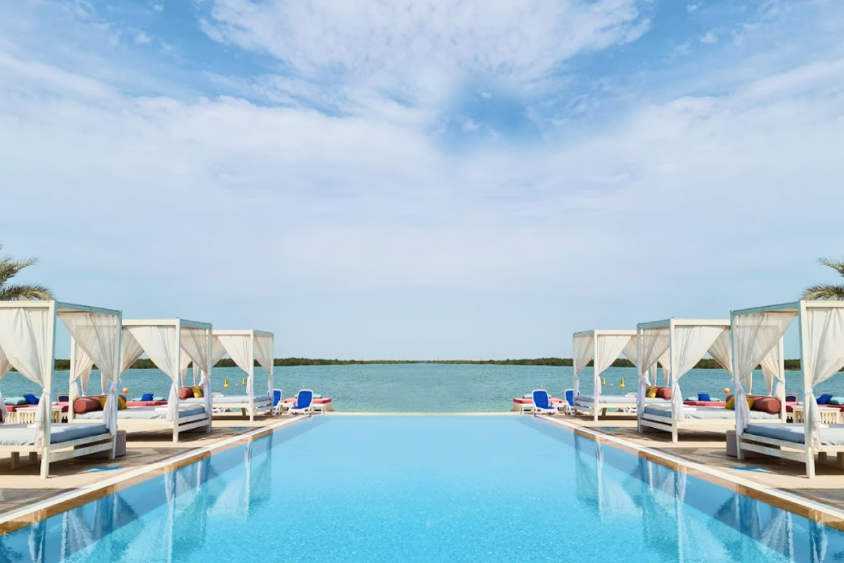 Yas Beach, Abu Dhabi A serene infinity pool with lounge beds and canopies overlooking calm waters and a blue sky with wispy clouds.