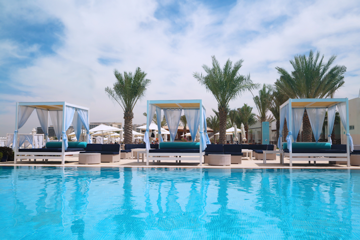 An inviting pool area featuring lounge beds with canopies, surrounded by palm trees under a bright blue sky.
