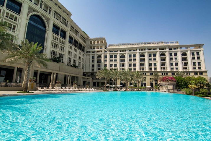 Hotel pool with clear water, surrounded by palm trees and a large building in the background.