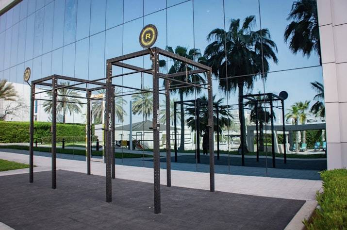 Outdoor fitness equipment with pull-up bars in front of a glass building reflecting palm trees.