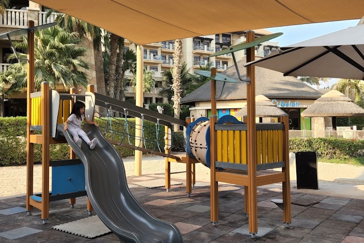 A child sliding down a gray slide on a colorful playground with climbing features and a shaded area