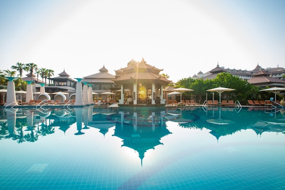 Pool with a reflective surface, surrounded by resort buildings and palm trees, set against a sunny sky