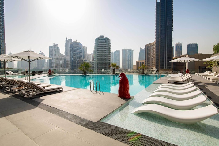 A person in a red outfit standing by a luxury pool with white lounge chairs, surrounded by tall buildings in a cityscape.