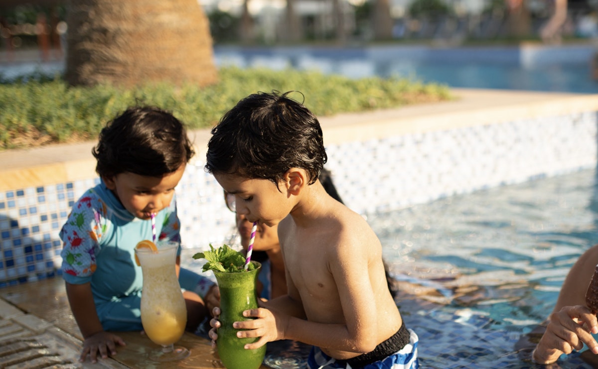Two children enjoying drinks by the pool, with one holding a green drink and the other a yellow one.