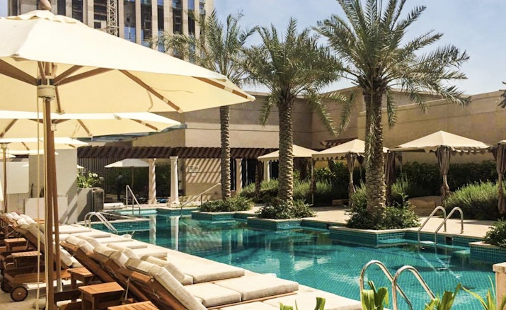 Hilton Al Habtoor City A serene poolside view with lounge chairs under umbrellas, surrounded by palm trees and a city building in the background.