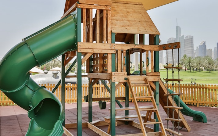Address Montgomerie A children's wooden playground with a green slide and various climbing features, with a city skyline in the background.