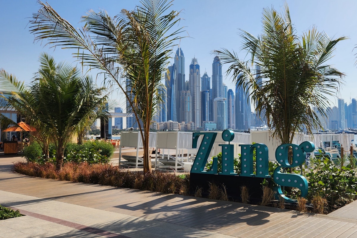 A sign reading "Zing" surrounded by greenery, with a skyline of tall buildings in the background.