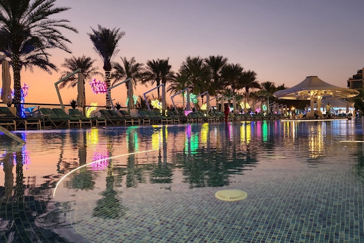 A vibrant pool area at dusk, illuminated by colorful lights and surrounded by palm trees.