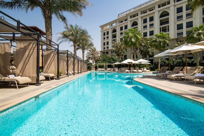 A luxurious pool surrounded by palm trees and sun loungers, with a clear blue sky and a hotel in the background
