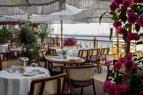 Chic outdoor dining area with woven chairs, fringe umbrellas, and vibrant flowers overlooking the water