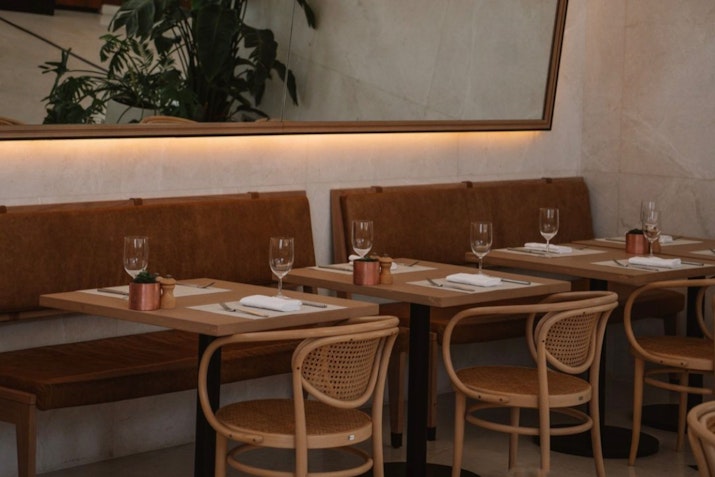 Interior of a restaurant with wooden chairs and tables set for dining, featuring plants and a mirror