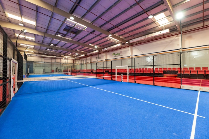 Indoor padel court with blue flooring and red seating.