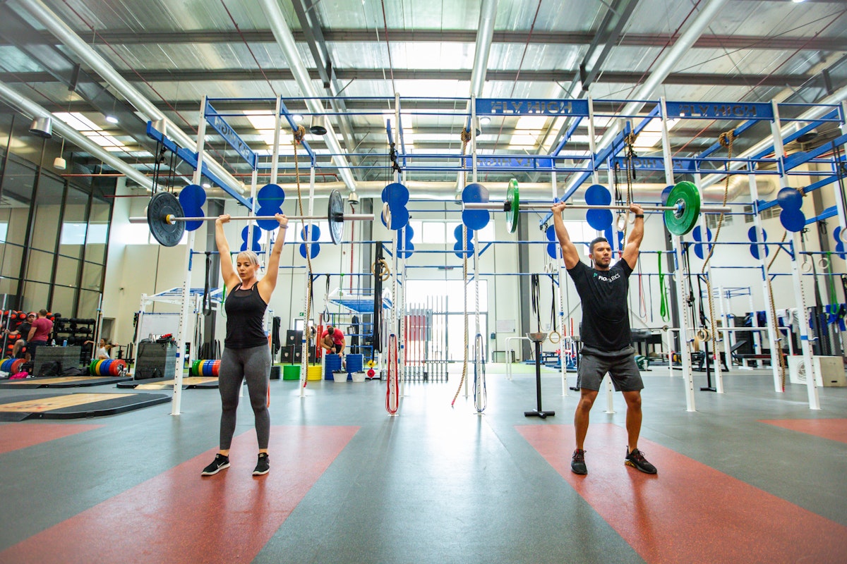 Two people lifting weights in a spacious, well-lit gym with high ceilings and various fitness equipment.