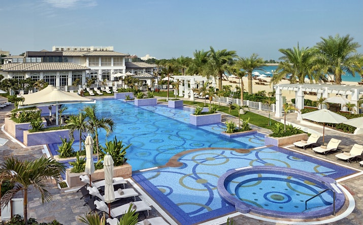 A scenic view of a resort pool area with patterned tiles, surrounded by palm trees and sun loungers.