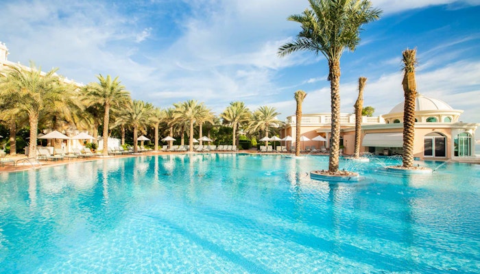 Kempinski Hotel & Residences A luxurious pool surrounded by palm trees and elegant lounge chairs, with a bright sky and resort-style architecture in the background.