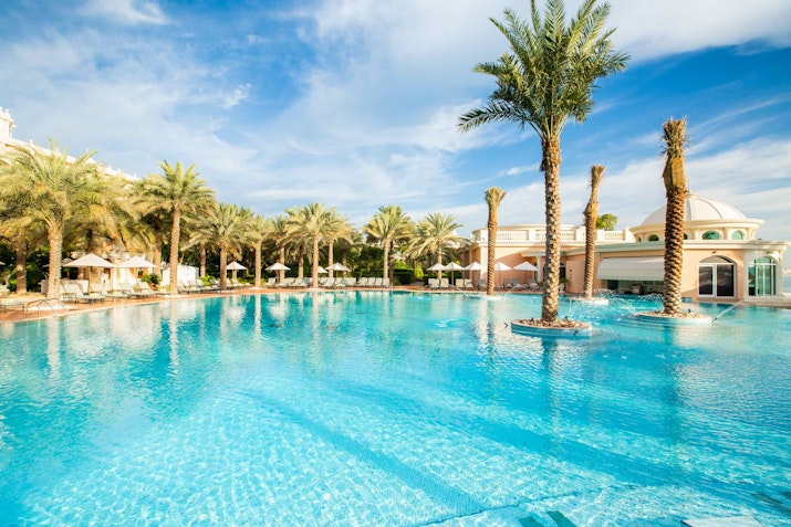 A luxurious pool surrounded by palm trees and elegant lounge chairs, with a bright sky and resort-style architecture in the background.