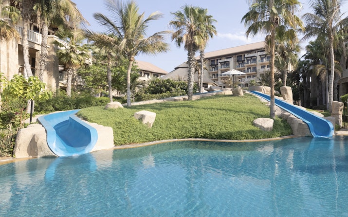 A beautiful pool with two water slides and surrounded by lush greenery and palm trees, with a resort building in the background.
