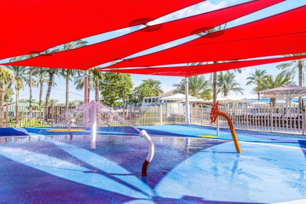 Splash pad with colorful water features under a red shade, surrounded by palm trees.