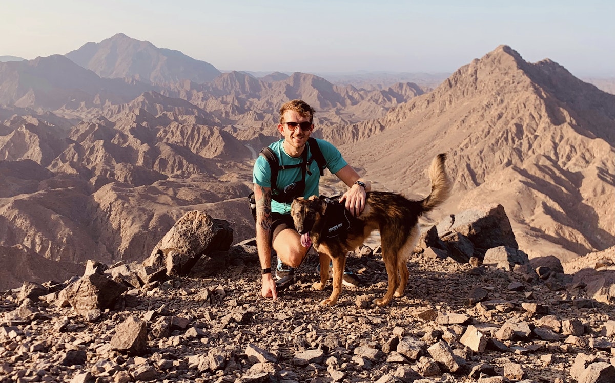 Man and dog posing on a mountain top with a scenic backdrop.