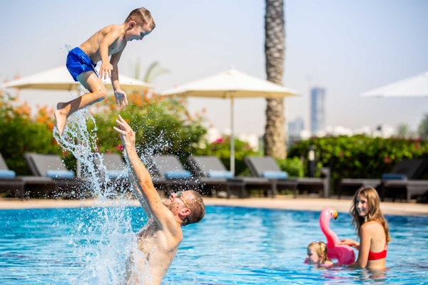 A father catching his son who is being thrown in the air above the water, while a mother and daughter play in the background of a sunny swimming pool.