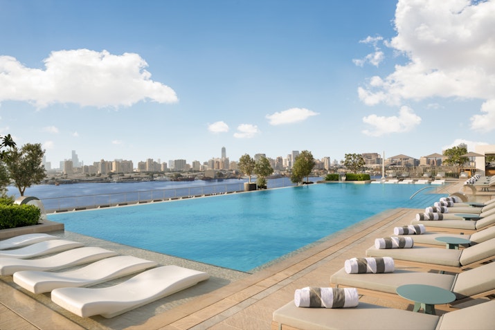 Elegant rooftop infinity pool with in-water loungers, overlooking a city skyline and river under a partly cloudy sky