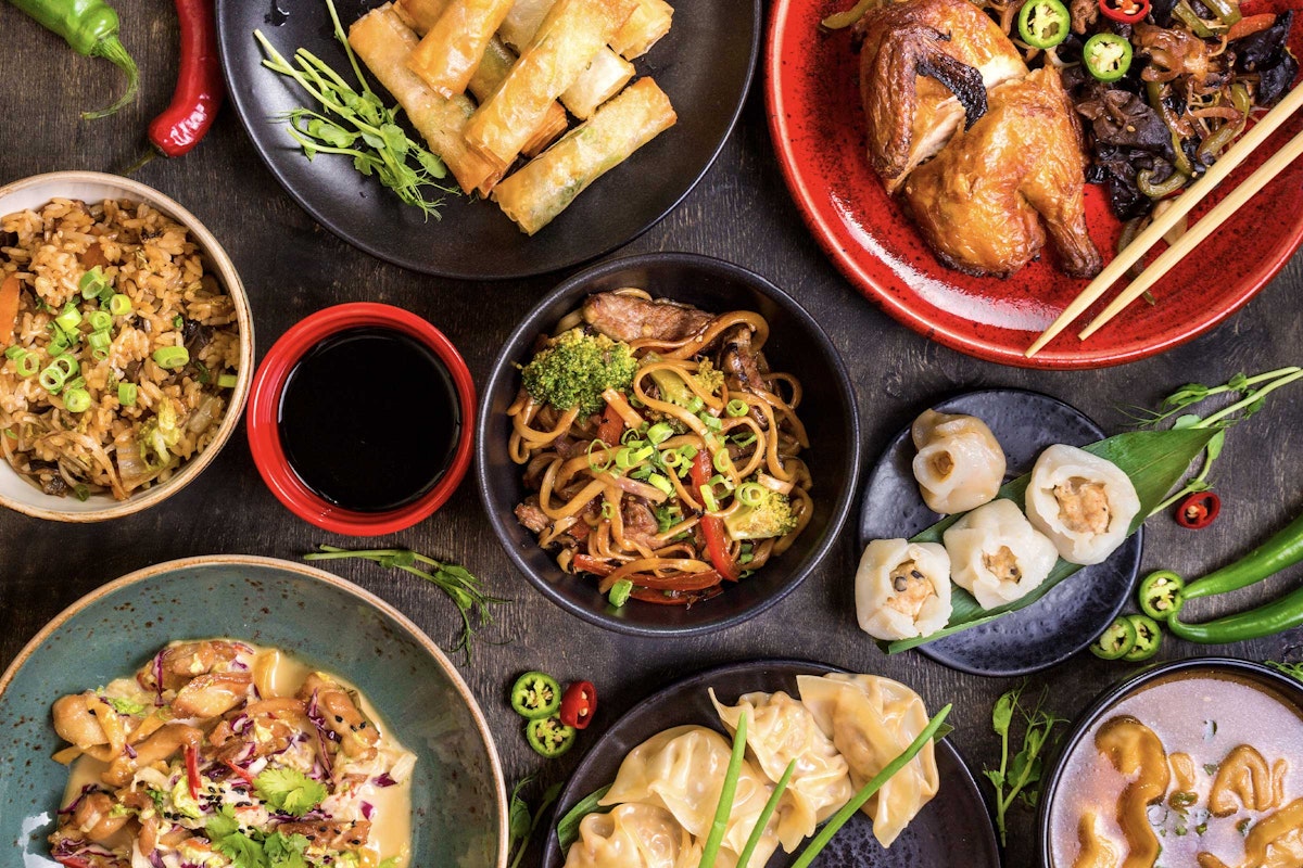A colorful assortment of Asian dishes, including noodles, dumplings, spring rolls, fried rice, and roasted chicken, displayed in vibrant bowls and plates.