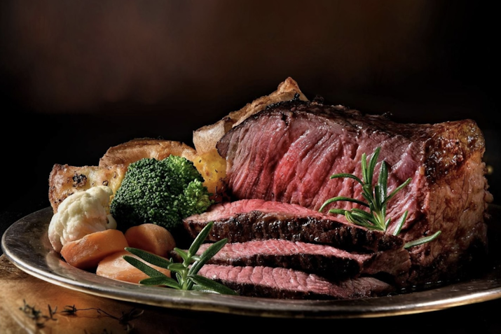 Close-up of a perfectly cooked roast beef with sides of roasted potatoes, broccoli, and carrots, garnished with fresh rosemary.