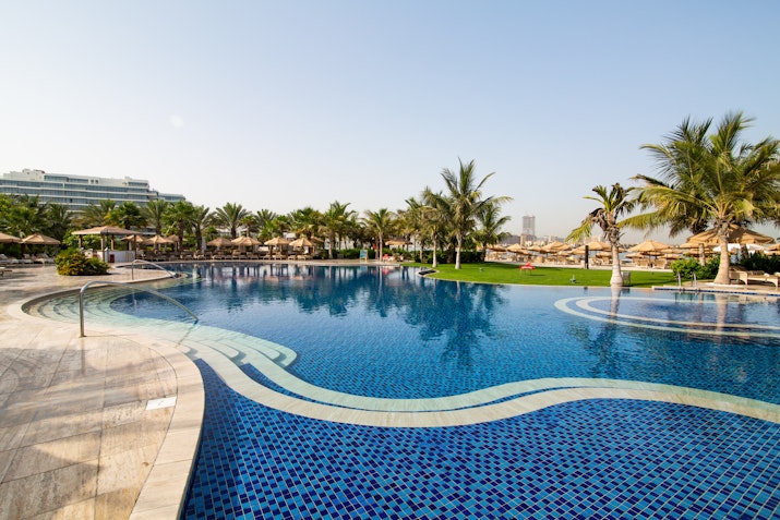 Waldorf Astoria Palm Jumeirah A large blue pool with a curved design, surrounded by palm trees and lounge chairs, under a clear sky.