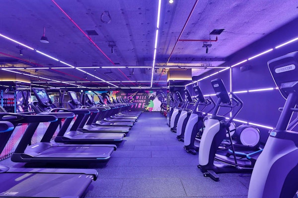 A modern gym interior featuring several treadmills lined up under vibrant purple and pink neon lights.