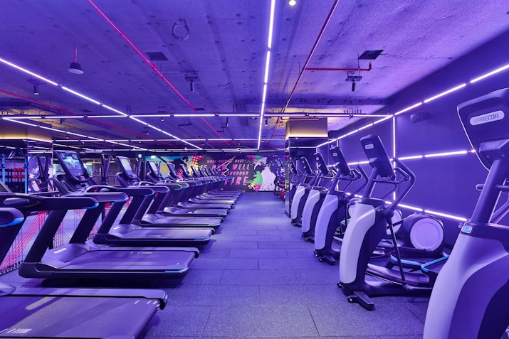 A modern gym interior featuring several treadmills lined up under vibrant purple and pink neon lights.