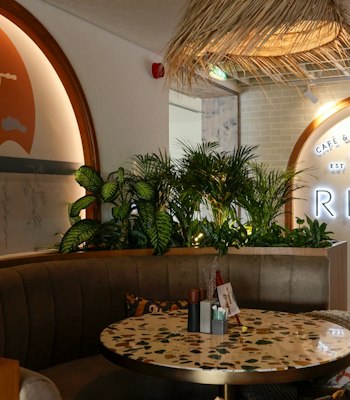 Experience the cozy ambiance at Risen Café & Artisanal Bakery. A stylish seating area, featuring greenery, a mural, and warm lighting.